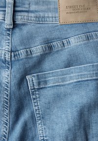 Jeans de mezclilla azul claro con una textura suave, que presentan un bolsillo trasero y una etiqueta de cuero grabada con "Street One Denim Atelier."