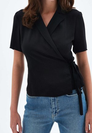 Femme portant un chemisier noir à manches courtes, noué sur le côté, associé à un jean denim bleu sur un fond uni.