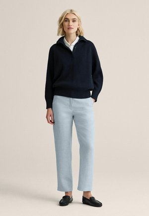 Kvinde stående iført en mørkeblå ribbet sweater over en hvid skjorte med krave, lyseblå bukser og sorte slip-on sko foran en beige baggrund.