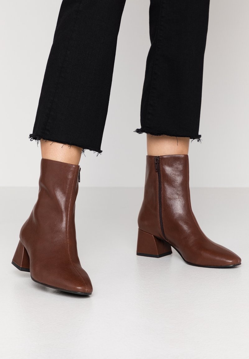 Jambes portant un pantalon noir raccourci et des bottines en cuir marron à talons carrés avec des fermetures éclair latérales sur un sol clair.