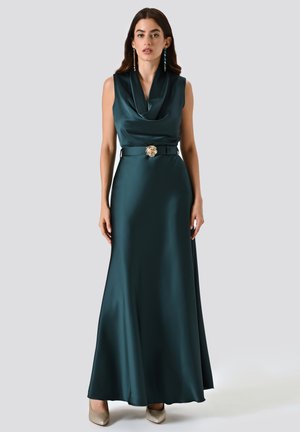 Donna che indossa un abito senza maniche in satin verde scuro con scollatura a cowl, fibbia dorata in vita, orecchini statement e tacchi dorati luccicanti su uno sfondo semplice.