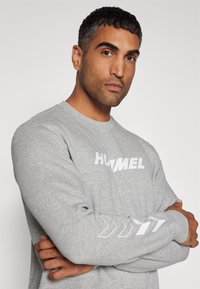 Grijze sweatshirt met lange mouwen, voorzien van een witte "HUMMEL"-logo aan de voorkant en een richtingspatroon op de linkermouw. Zachte textuur.