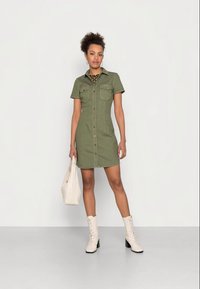 Vestido de manga curta, verde-azeitona, com fecho de botão à frente, dois bolsos no peito e uma silhueta recta. Combinado com botas de combate em creme.