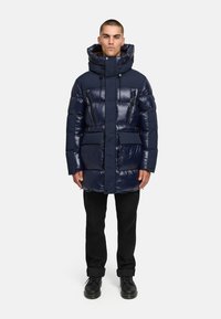 Laulukavojeg tamnoplave jakne s visokom ovratnikom, zatvorenim džepovima i sjajnom završnom obradom. Debela podstava osigurava izolaciju. Nosena je preko crnih hlača i cipela.