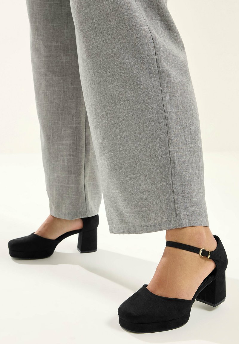 Osoba w szarych szerokich spodniach i czarnych platformach na grubym obcasie z paskami na kostce, stojąca na jasnej powierzchni.