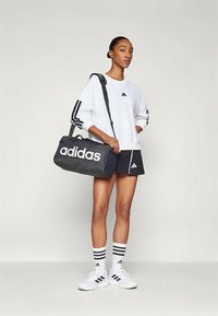 Γυναίκα με λευκή μπλούζα Adidas, μαύρες βερμούδες Adidas, λευκές κάλτσες και παπούτσια Adidas, κρατώντας μια μαύρη τσάντα αθλητισμού Adidas στον ώμο.