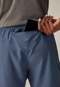 Kék sportpantalon, zippzáros zsebbel, amely egy okostelefont tartalmaz. Simogatós, könnyű anyagból készült, rugalmas derékpánttal.