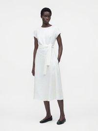 Femme debout portant une robe midi blanche à manches courtes avec une ceinture à la taille, associée à des chaussures plates noires, sur un fond blanc uni.