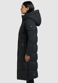 Lang zwarte donzen jas met een aansluitende capuchon, geborduurd ontwerp, zijzakken en logopatch op de mouw. Glad textuur en midi-lengte.