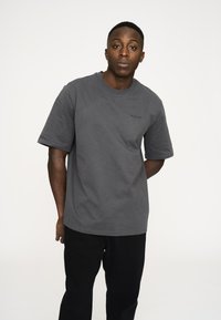 Grå bomulds-t-shirt med korte ærmer, rund halslinje og diskret branding på brystet. Sammen med sorte bukser poserer personen afslappet.