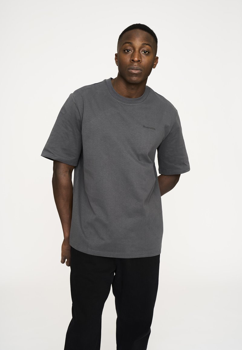 Grå bomulds-t-shirt med korte ærmer, rund halslinje og diskret branding på brystet. Sammen med sorte bukser poserer personen afslappet.