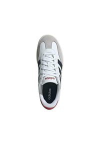 Scarpa sportiva bianca con dettagli in suede grigio, strisce blu navy e branding rosso. Presenta una chiusura con lacci e una suola in gomma testurizzata.