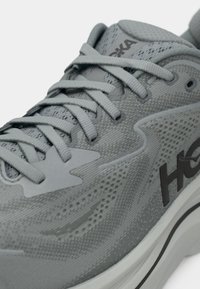 Close-up de um sapato atlético cinza com tecido em malha, atacadores a condizer e logótipo "Hoka" na lateral, mostrando detalhes da textura e costura.