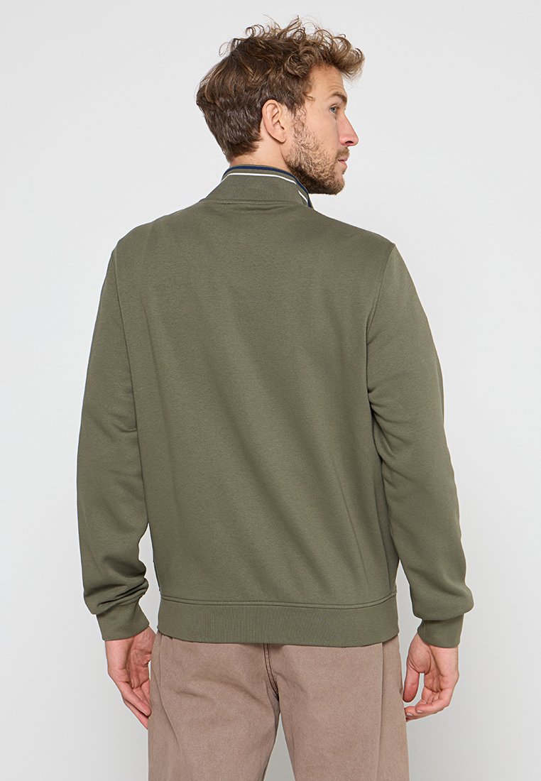 Uomo con capelli ricci che indossa una felpa verde oliva e pantaloni beige, in piedi con la schiena rivolta e la testa di profilo.