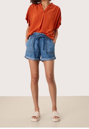 Blouse manches courtes couleur rouille avec col en V ; associée à un short en jean avec revers et deux poches avant. Sandales légères.