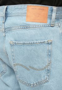 Primer plano de unos pantalones vaqueros de mezclilla azul claro, mostrando el bolsillo trasero y la etiqueta de cuero marrón con el nombre de la marca "JACK & JONES".