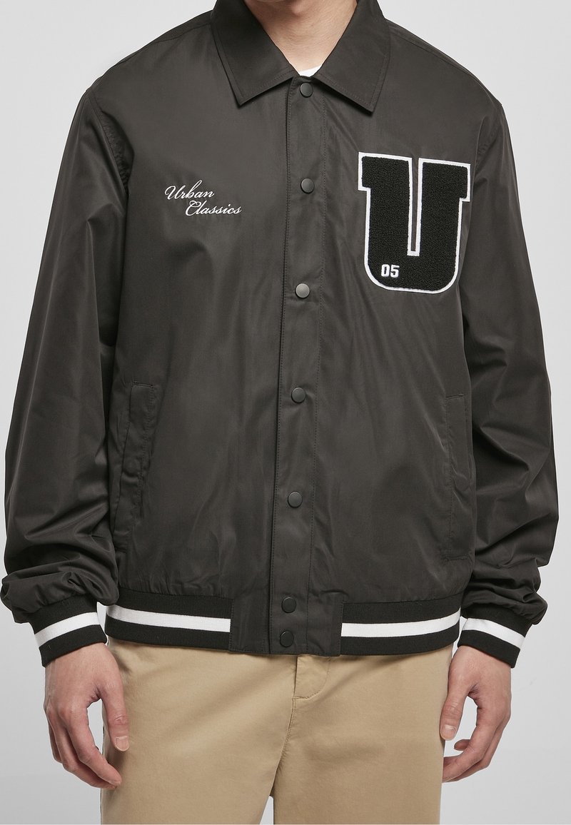 Veste noire à boutons avec texte blanc "Urban Classics" sur la poitrine gauche et grand patch noir et blanc "U 05" sur la poitrine droite, portée avec un pantalon beige.