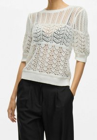 Maglione lavorato a maglia di colore bianco sporco con motivo in pizzo aperto, maniche corte, polsini e orlo a costine, abbinato a pantaloni neri. Design leggero e dalla texture interessante.