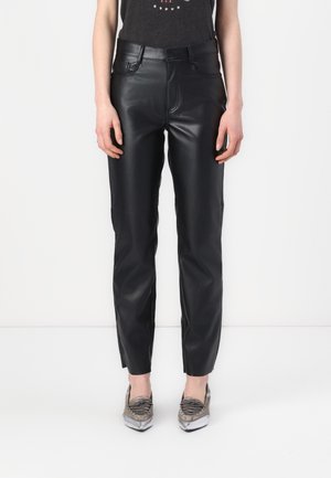 Femme portant un pantalon droit en similicuir noir et des mocassins métalliques à bout pointu sur fond blanc.
