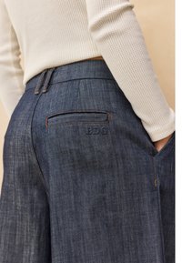 Pantalones de mezclilla anchos de tono oscuro con una textura sutil, que cuentan con un bolsillo trasero con un logo bordado y detalles de costura en contraste.