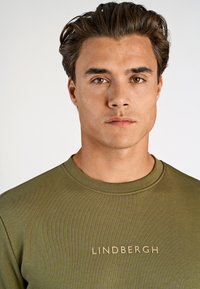 Koszulka typu crewneck w oliwkowym kolorze z wyhaftowanym logo "LINDBERGH" w jasnobeżowym odcieniu. Gładka tekstura i dopasowany krój.