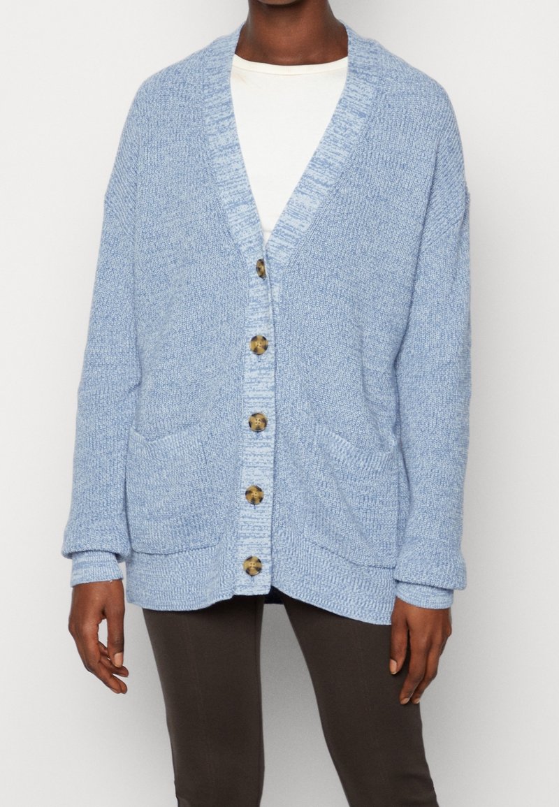 Personne portant un cardigan tricoté surdimensionné bleu clair avec des poches avant et des boutons marron par-dessus une chemise blanche et un pantalon foncé.