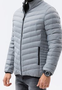 Szara kurtka puchowa o pikowanym wzorze, z wysokim kołnierzem i czarnymi zamkami błyskawicznymi. Posiada dwie boczne kieszenie i dopasowany krój, wykonana z lekkiego materiału.