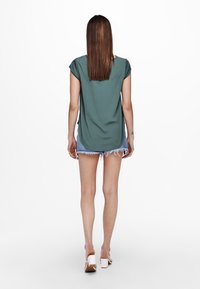 Top verde a maniche corte con una vestibilità rilassata, caratterizzato da un orlo arrotondato. Abbinato a pantaloni corti in denim chiaro e sandali bianchi con tacco a blocco.