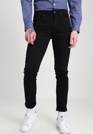 Personne portant un pantalon noir slim, une chemise bleue boutonnée, des baskets blanches Nike et des tatouages visibles sur les mains sur un fond clair.