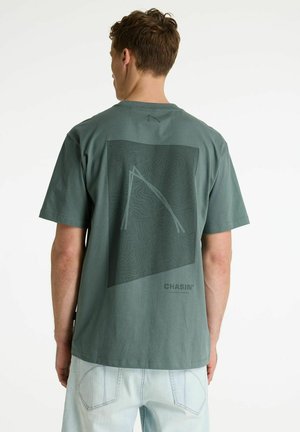 Man draagt een groen t-shirt met een topografische kaart en de tekst "CHASIN'" op de achterkant, gecombineerd met lichtblauwe jeans.