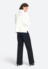 Cremiger Strickpullover mit hohem Kragen und seitlichen Schlitzen, kombiniert mit schwarzen, weit geschnittenen Hosen und schwarzen Absatzschuhen.