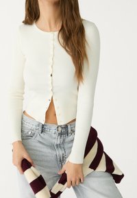 Ribbed cream long-sleeve cardigan with round neckline and scalloped button detail; paired with light denim jeans and a maroon striped sweater.
