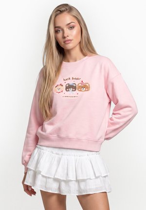 Junge Frau mit langen blonden Haaren trägt rosa Sweatshirt mit drei Comic-Bären und weißem Rüschenschwanz vor weißem Hintergrund.