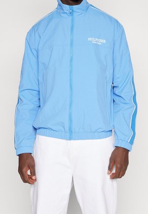 Man wearing light blue zip-up jacket with elastic cuffs and "HILFIGER NEW YORK" text on chest, paired with white pants.