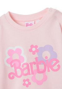 T-shirt en coton rose avec un col rond, arborant un grand logo "Barbie" et des motifs floraux colorés en rose, violet et blanc.