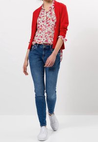 Un blazer rouge sur un blouse à fleurs, un jean bleu et des baskets blanches. La tenue présente un mélange de motifs et de couleurs vives.