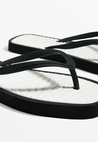 Zwarte rubberen flip-flops met dunne bandjes, een witte gestructureerde voetbed, en een zwarte zool. Heeft een diagonaal lijnpatroon op de binnenzool.