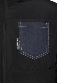 Czarna kurtka puchowa z ciemnym denimowym kieszonką z widocznym przeszyciem. Zamek błyskawiczny na środku. Na ramionach widoczna teksturowana tkanina.