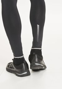 Schwarze sportliche Leggings mit einer eng anliegenden Passform, ausgestattet mit reflektierenden Streifen und einer glatten Textur. Kombiniert mit schwarzen Sneakers und Crew-Socken.
