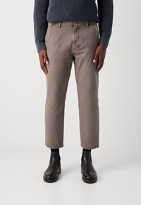 Pantalons cropped marron clair avec des poches visibles et des passants de ceinture, associés à des bottines noires. Le tissu semble lisse avec une texture minimale.