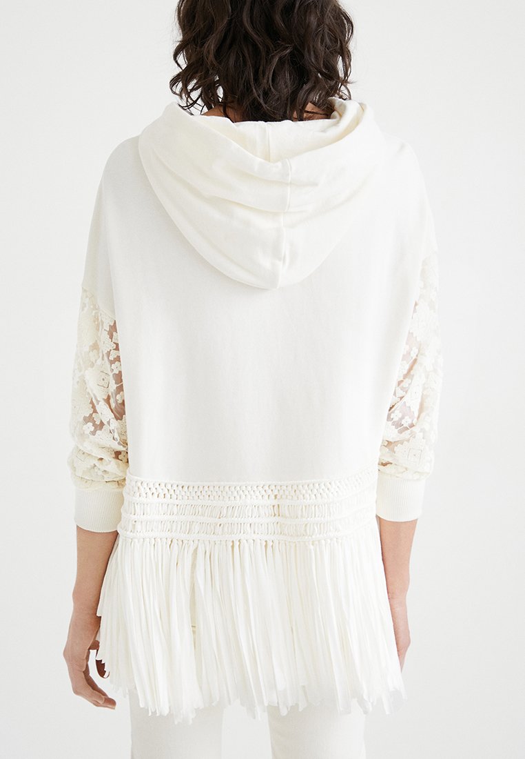 Felpa bianca con maniche in pizzo e orlo frangiato. Presenta una vestibilità comoda, cappuccio e vita arricciata. Tessuto morbido e testurizzato con dettagli intricati.