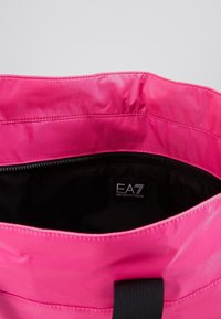 Růžová taška přes rameno s hladkým povrchem, černým vnitřkem a zapínáním na zip. Má plochý popruh a uvnitř štítek s logem.