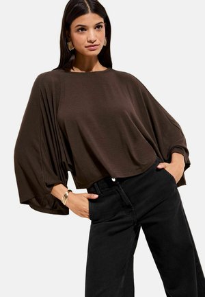 Brūns, vaļīgs krekls ar platiem piedurknēm, kombinēts ar augsta vidukļa melnām džinsām. Materiāls šķiet mīksts; dizains ir atvieglots un brīvi krītošs.