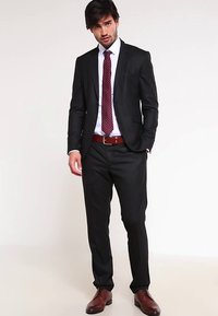 Schwarzer Anzug mit tailliertem Schnitt, weißem Hemd und bordeauxroter Krawatte. Brauner Ledergürtel und passende Schuhe vervollständigen das Outfit.