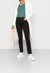 Vit lång kappa över en turkos kabelstickad tröja, svarta byxor och vita plattformssneakers. Håller en ljusrosa och blå väska.