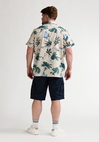 Chemise à manches courtes beige à motifs floraux verts et bleus, associée à un short cargo bleu marine, des baskets blanches et des chaussettes rayées.
