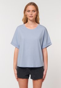 A young woman stands against a neutral background, wearing a light blue oversized t-shirt and black shorts, with a neutral expression.