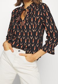 Blusa nera con motivo zigzag rosso e arancione, presenta un collo a cravattino con nappine, maniche lunghe con polsini a ruches, abbinata a pantaloni bianchi.