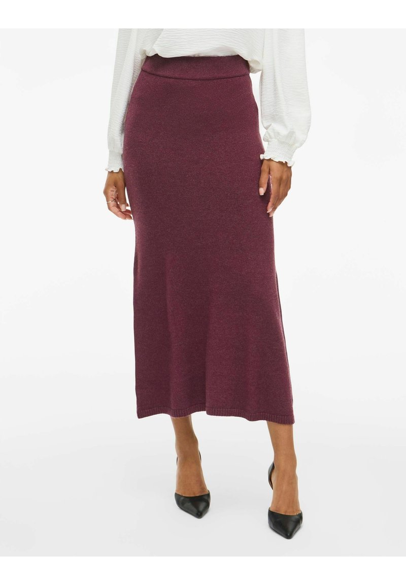 Femme portant une jupe en maille bordeaux taille haute, longueur cheville, un chemisier blanc à manches longues et des talons noirs à bout pointu.