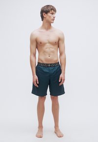 Ανδρικό μαγιό σε σκούρο τιρκουάζ χρώμα με μαύρη ζώνη με σχέδια, με λεία υφή και μήκος μέχρι το γόνατο. Ξυπόλητος, στέκεται όρθιος.